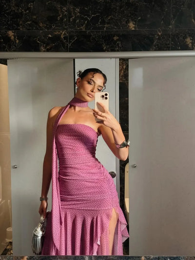 Woman in pink polka-dotted asymmetric ruffle dress with thigh-high slit and scarf, posing in a marble bathroom.