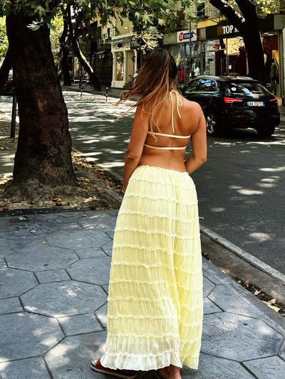 Back view of a woman in a pale yellow, tiered ruffle halter co-ord set with neck and back ties, strolling city street.