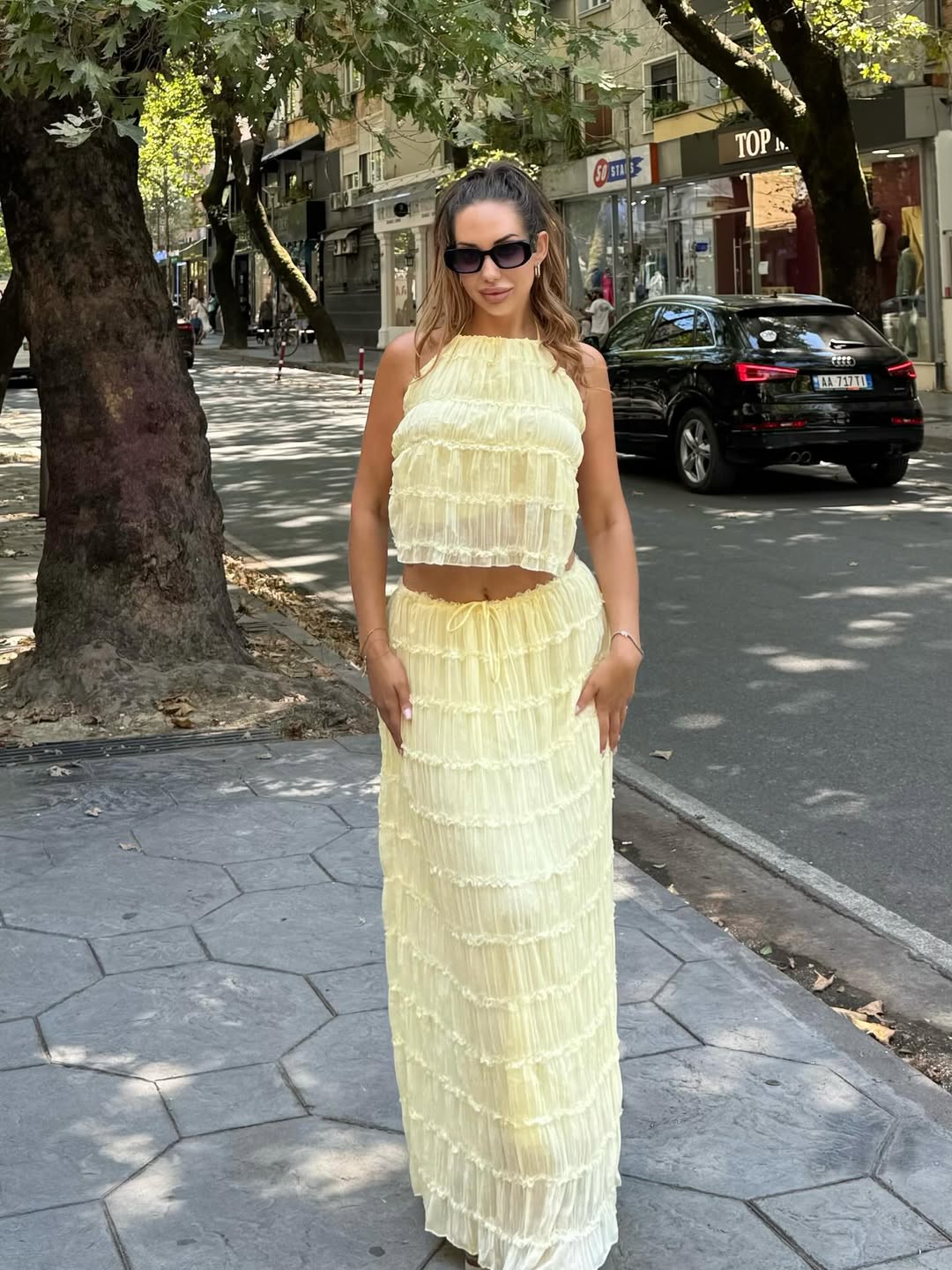 Woman wearing pale yellow cloud-ruffle halter crop top and matching tiered maxi skirt, sunglasses, on a city sidewalk.