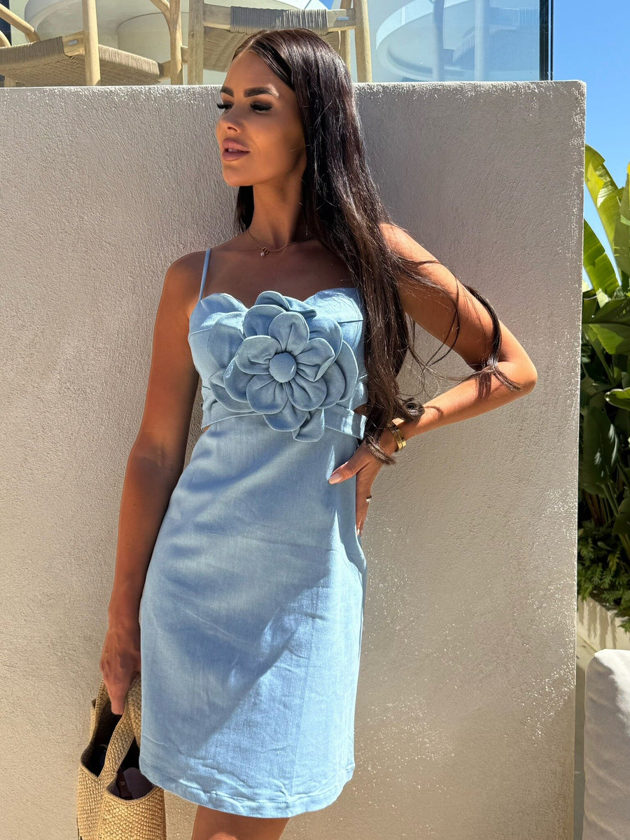 Light blue spaghetti-strap dress with a large layered fabric flower applique at the bodice; model holds a straw tote.
