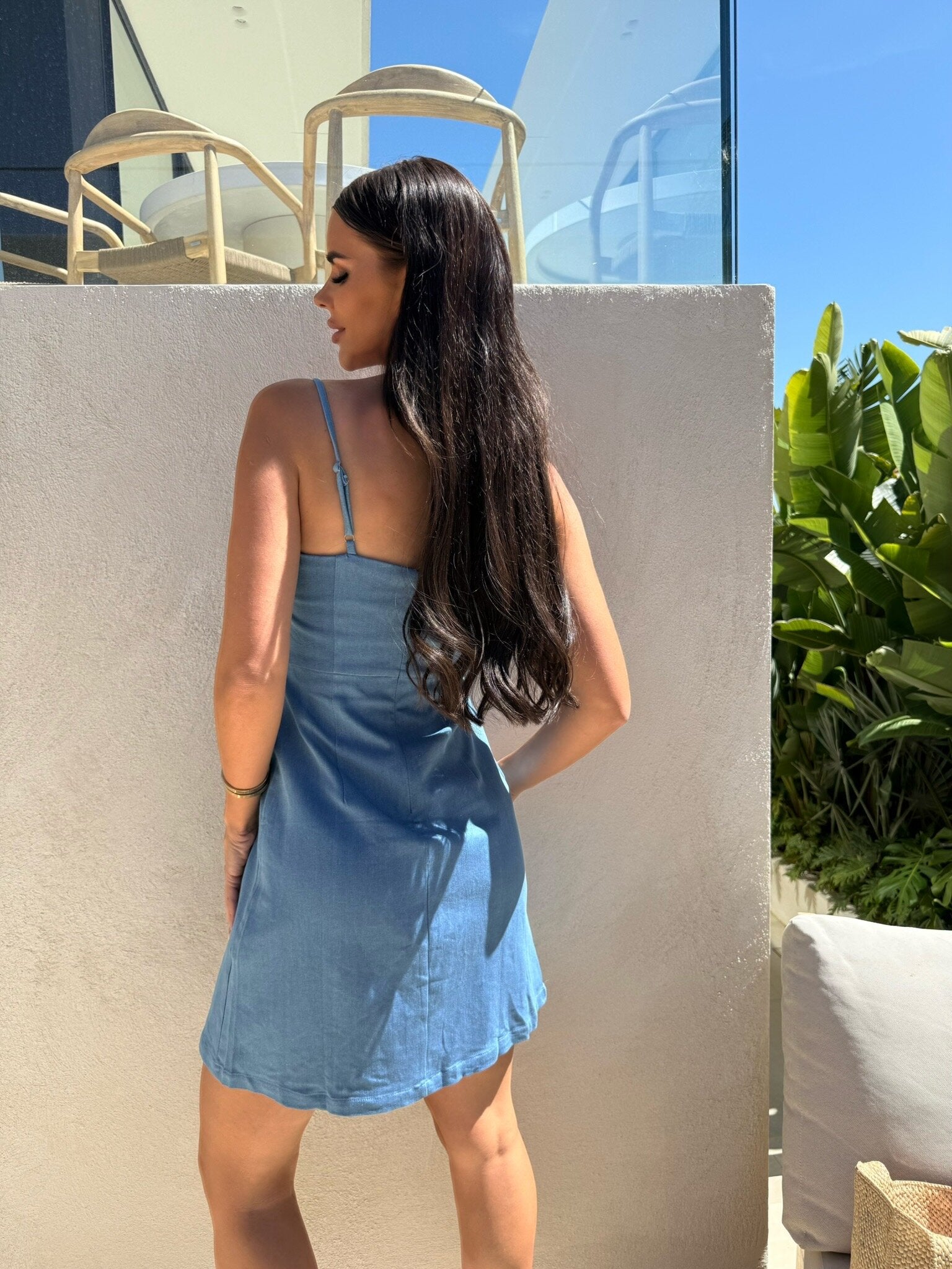 Woman in light blue spaghetti-strap dress, back view, A-line mini, outdoors beside a white wall.