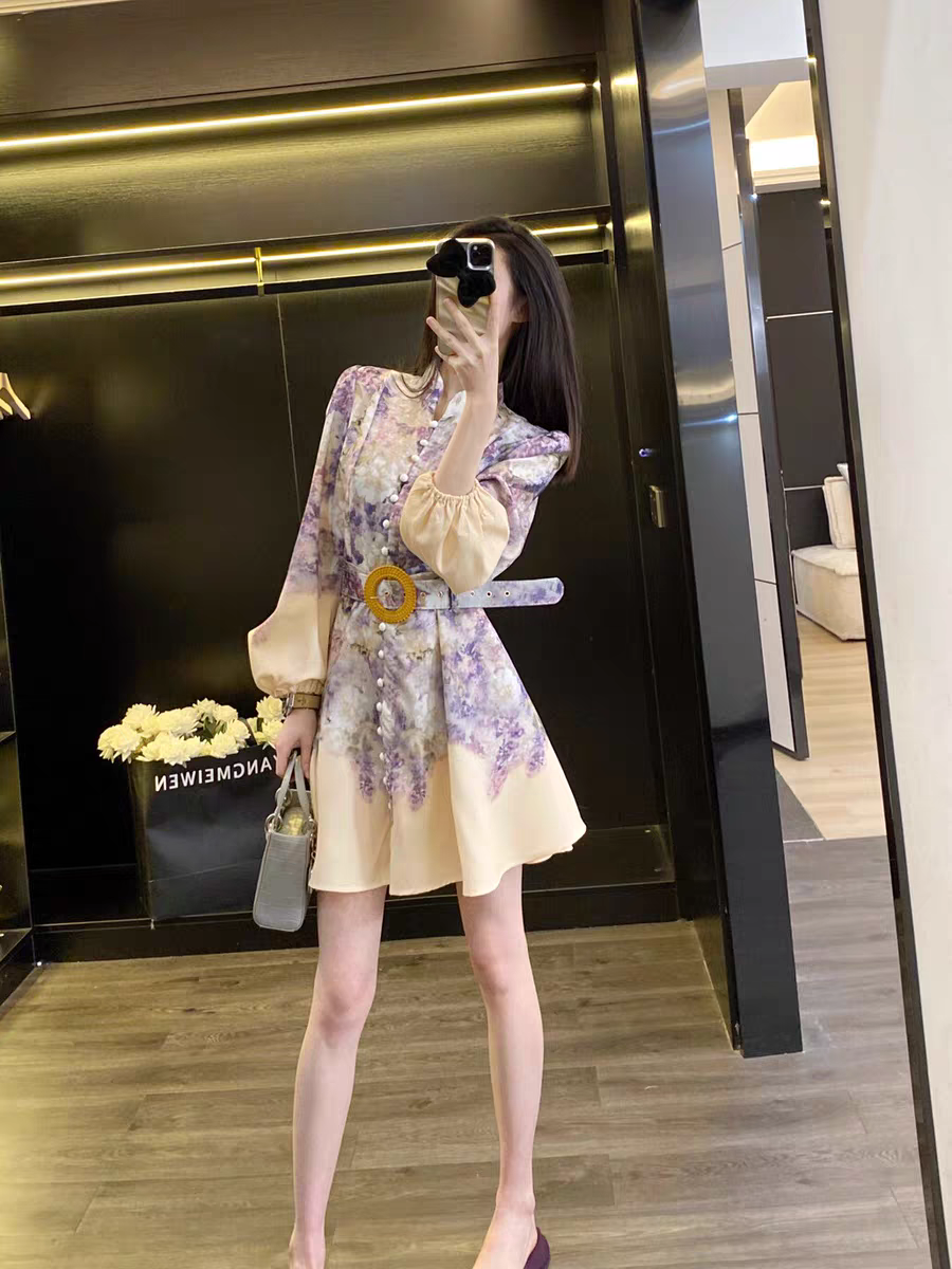 Woman posing in a mirror wearing a cream floral short A-line dress with beige puff sleeves and a yellow belt buckle, carrying a gray handbag.