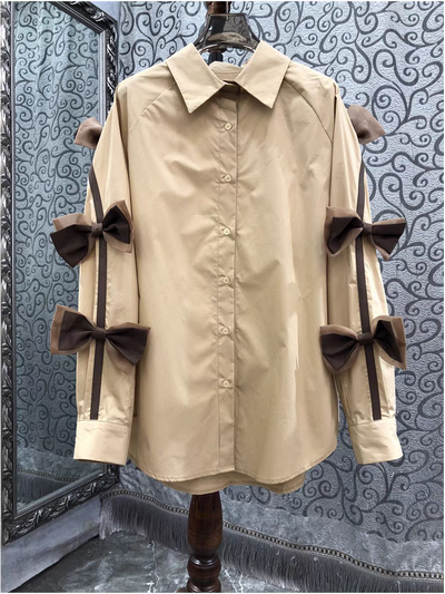 Beige oversized button-down shirt on a hanger with dark brown bow appliques along both sleeves, attached to black ribbons.