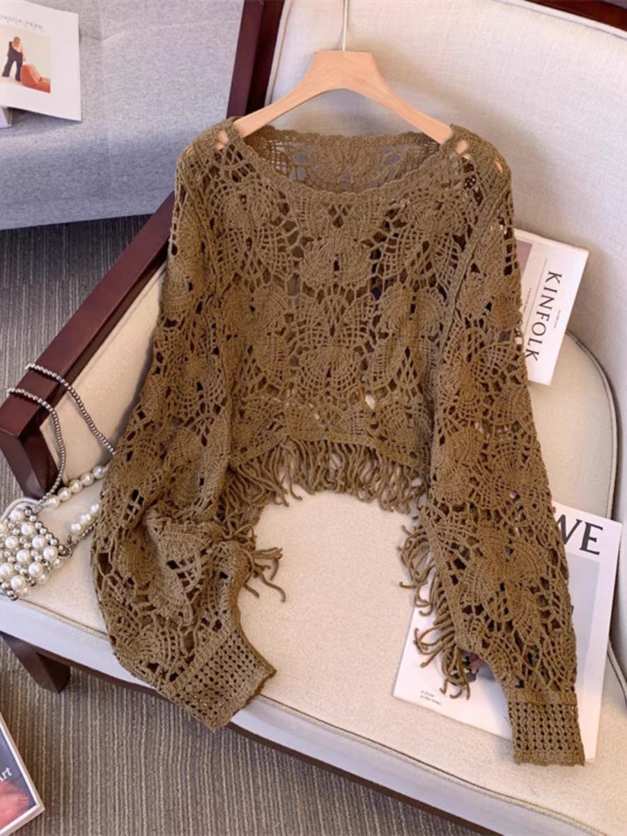 Taupe butterfly-cutout crochet top with fringe hem and long sleeves, hung on a wooden hanger over a beige chair.