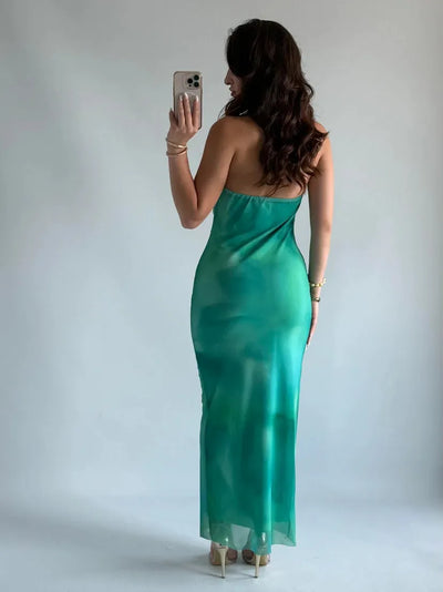 Back view of a woman in teal-to-green ombre halter-neck maxi dress with a draped cowl neckline, standing in a studio.