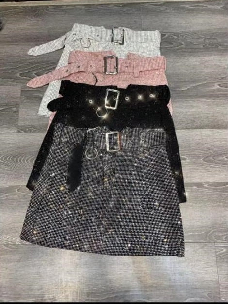 Row of rhinestone mini skirts in silver, pink, black, and gunmetal, each with metal belt and buckle on gray wood floor.