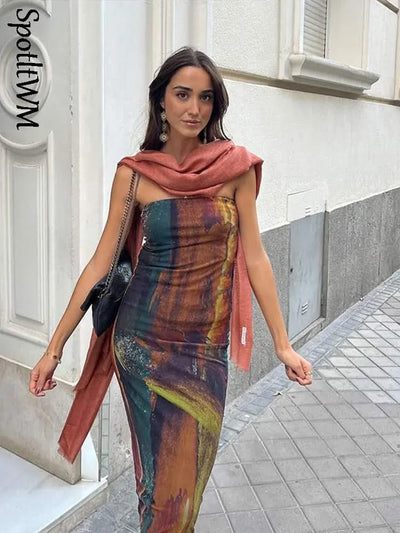 Woman in multicolored tie-dye mesh tube dress with coral scarf draped over shoulders, black crossbody bag on city sidewalk.