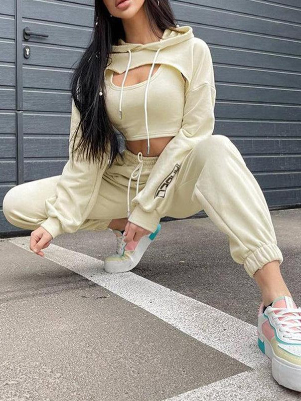 Beige cropped hoodie with cut-out chest and hood, paired with matching joggers and pastel sneakers, against a gray wall.