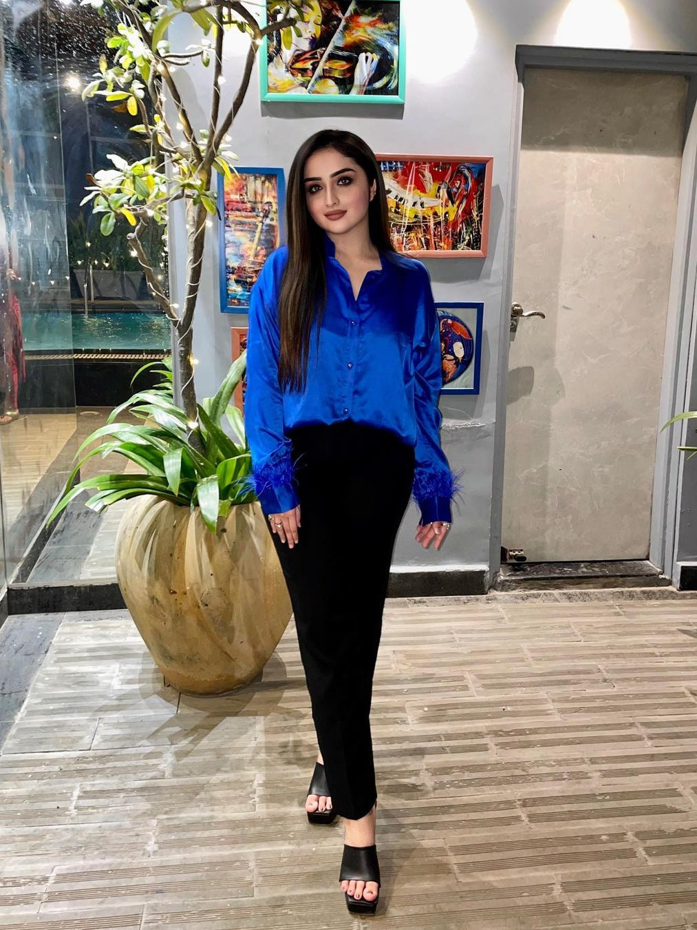 Woman in royal blue satin shirt with feathered sleeve cuffs, black pants, black heeled sandals, indoor gallery setting.