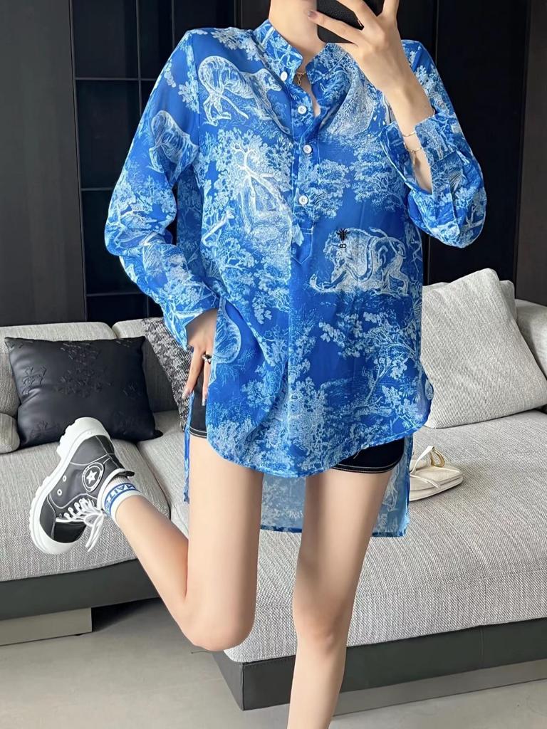 Blue oversized button-down shirt with white floral and animal print, curved hem, worn over black shorts indoors.