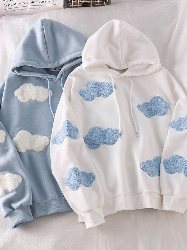 Two pastel hoodies on a bed: one light blue with white fluffy cloud patches, the other white with light blue cloud patches; both have drawstrings and ribbed cuffs.