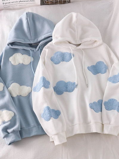 Two pastel hoodies on a bed: one light blue with white fluffy cloud patches, the other white with light blue cloud patches; both have drawstrings and ribbed cuffs.