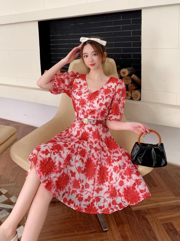 Woman in red floral A-line dress with puff sleeves and belt, white bow headband, sits on beige chair, black handbag.