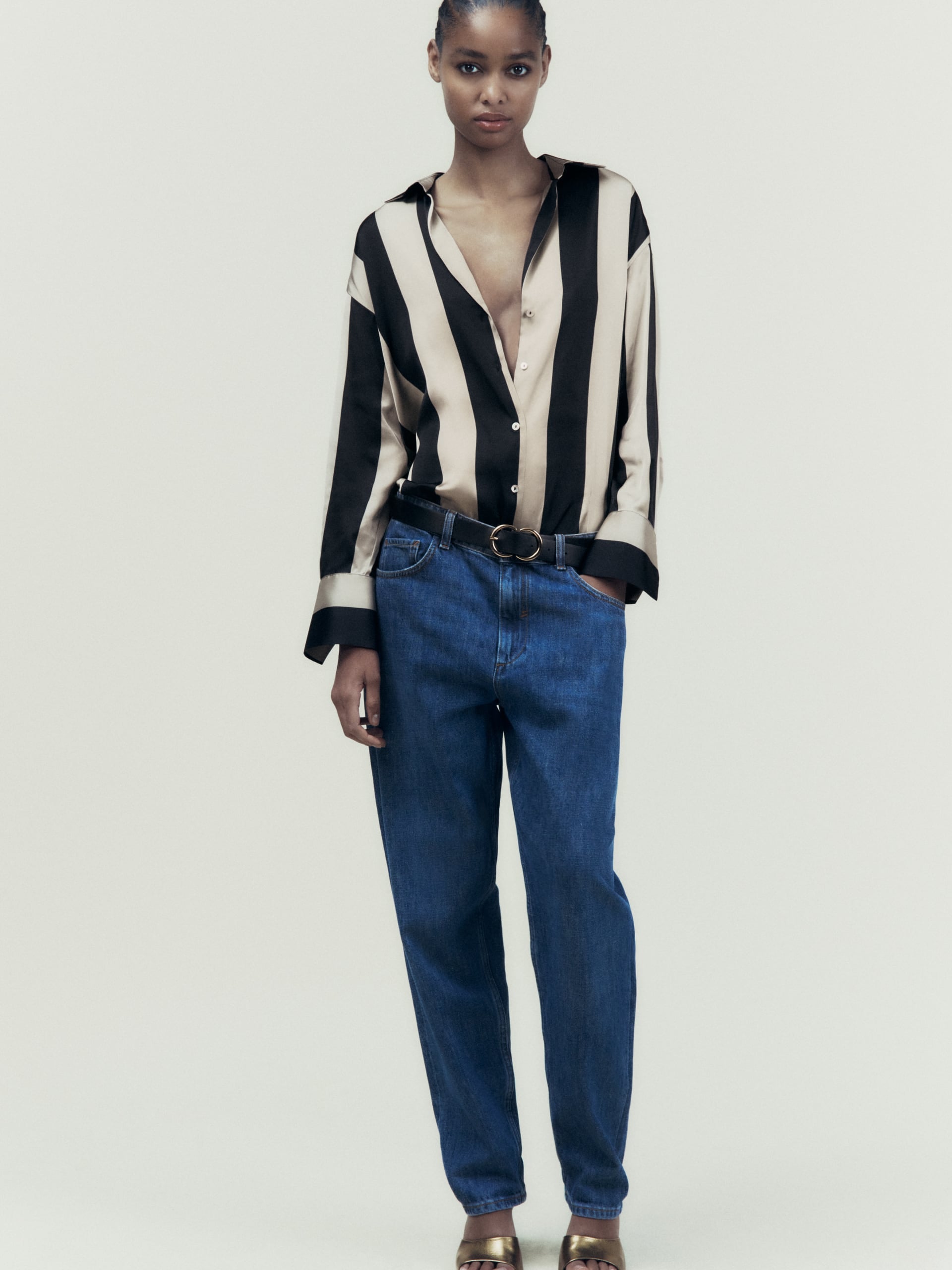 Model in beige and black striped satin shirt with drop shoulders, tucked into blue jeans with a double-ring belt.