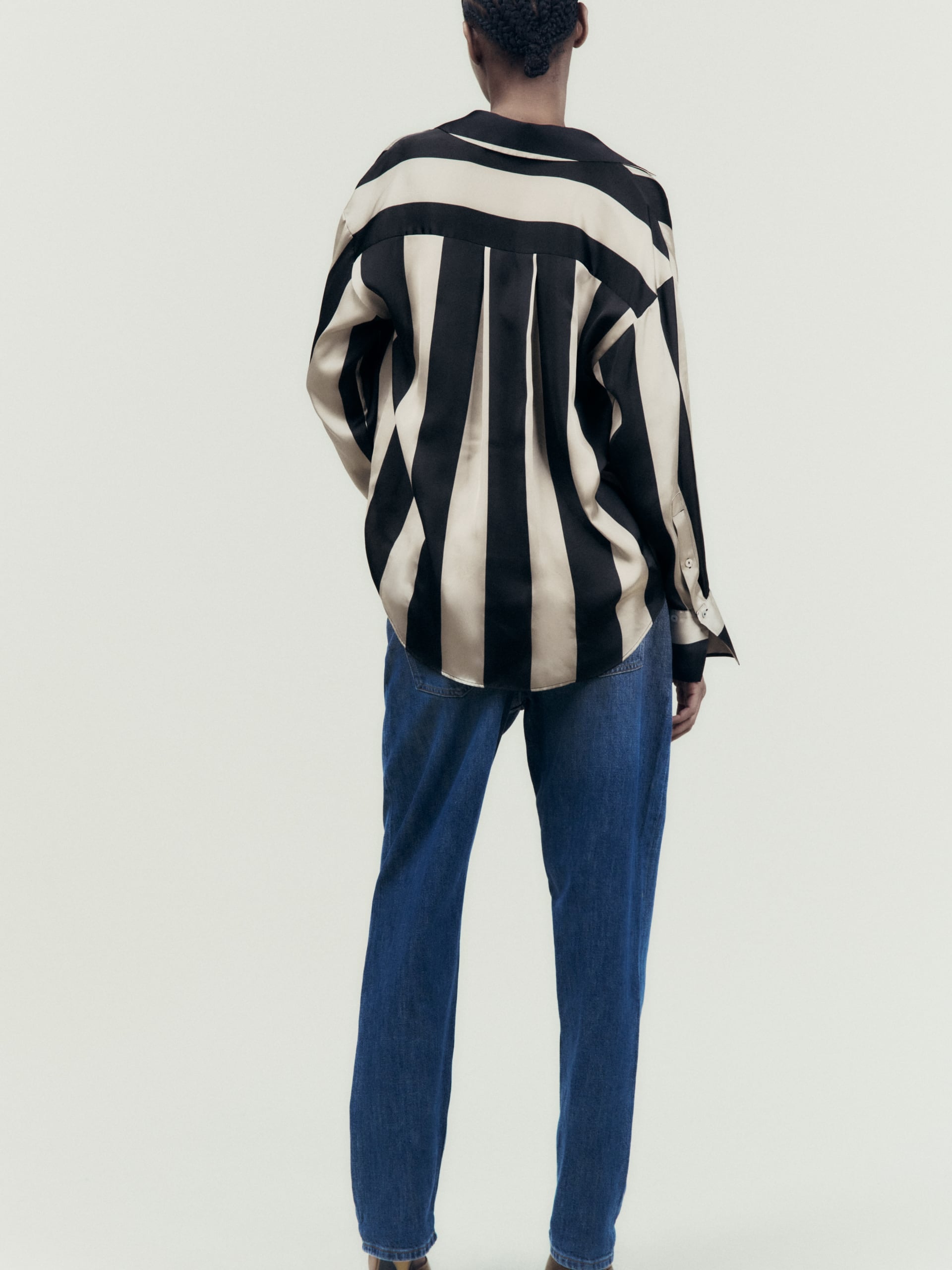 Back view of oversized satin shirt with beige and black stripes and drop-shoulder silhouette, paired with blue jeans.