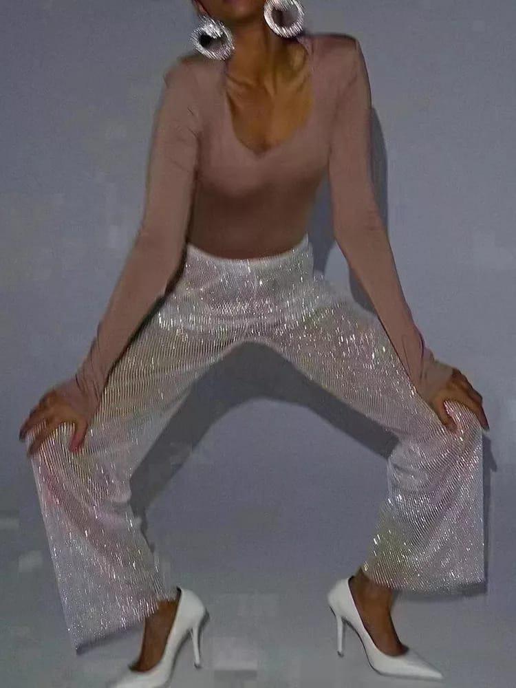 Model wearing sparkling rhinestone-studded silver pants, nude long-sleeve top, large hoop earrings, and white heels.