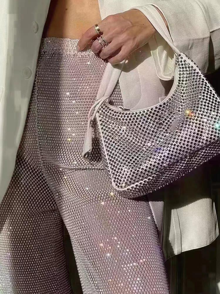 Rose-gold sequined pants with rhinestones and a crystal-studded white handbag held by a hand wearing rings.