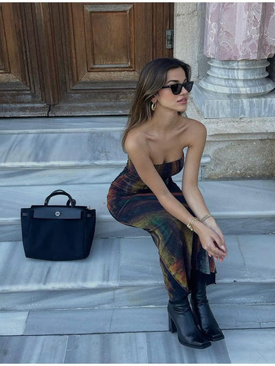 Woman in a strapless tie-dye mesh tube dress sits on marble steps with a black handbag; wearing sunglasses and boots.