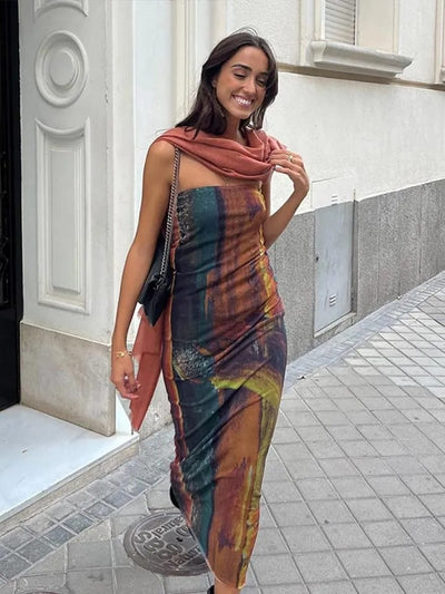 Smiling woman in a multicolor tie-dye mesh tube dress, rust scarf draped over shoulders, black chain-strap bag on a city sidewalk.