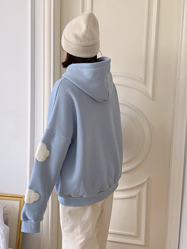 Back view of a light blue oversized hoodie with cloud patches on the sleeve, beige pants, and a white beanie.