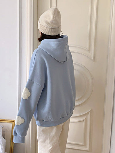 Back view of a light blue oversized hoodie with cloud patches on the sleeve, beige pants, and a white beanie.