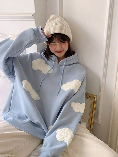 Light blue oversized hoodie with white fluffy cloud patches, drawstrings, worn by smiling young woman in beige beanie.