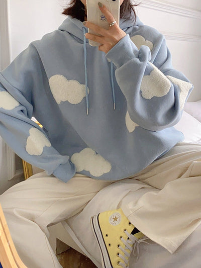Oversized light-blue hoodie with white cloud patches on sleeves and chest, blue drawstrings, worn with beige pants.