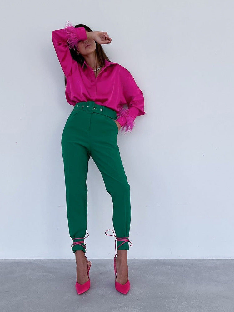 Model wearing pink satin shirt with feathered cuffs, green high-waisted pants with belt, and pink lace-up heels.