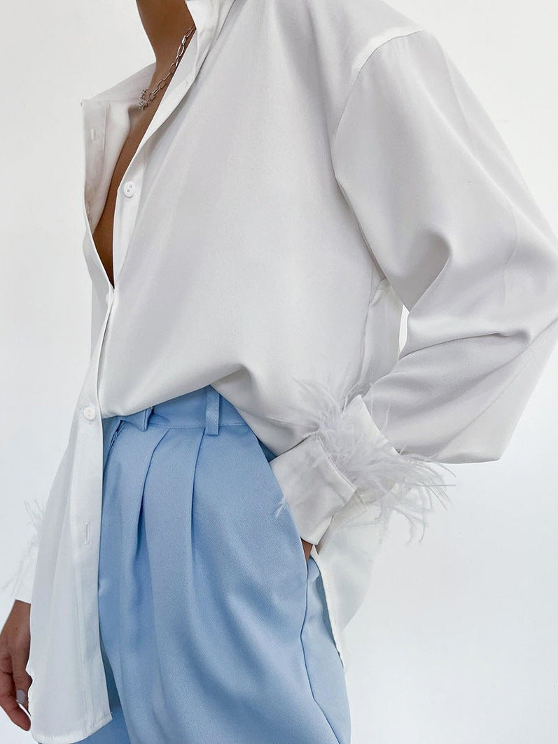 White satin shirt with feather-trimmed cuffs and an open neckline, paired with light blue pleated trousers.