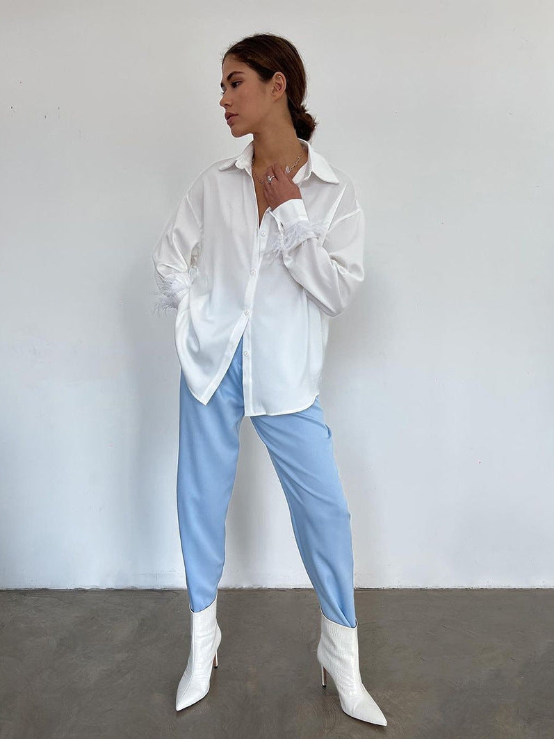 Woman in oversized white satin shirt with feather-trim sleeves, light blue pants, and white pointed-toe ankle boots.