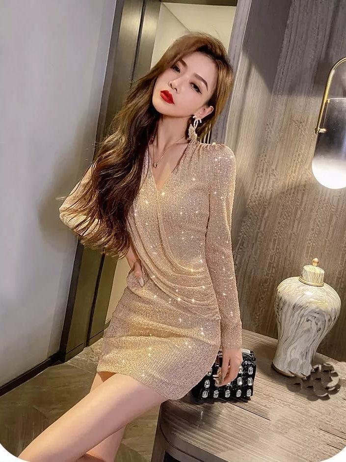 Woman in champagne-gold sequin mini dress with long sleeves and wrap detailing, posing with a black studded clutch.