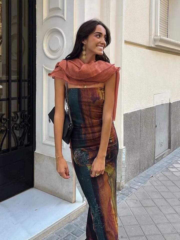 Woman wearing a multicolor tie-dye mesh tube dress with a rust scarf, gold earrings, and a black crossbody bag on a city sidewalk.