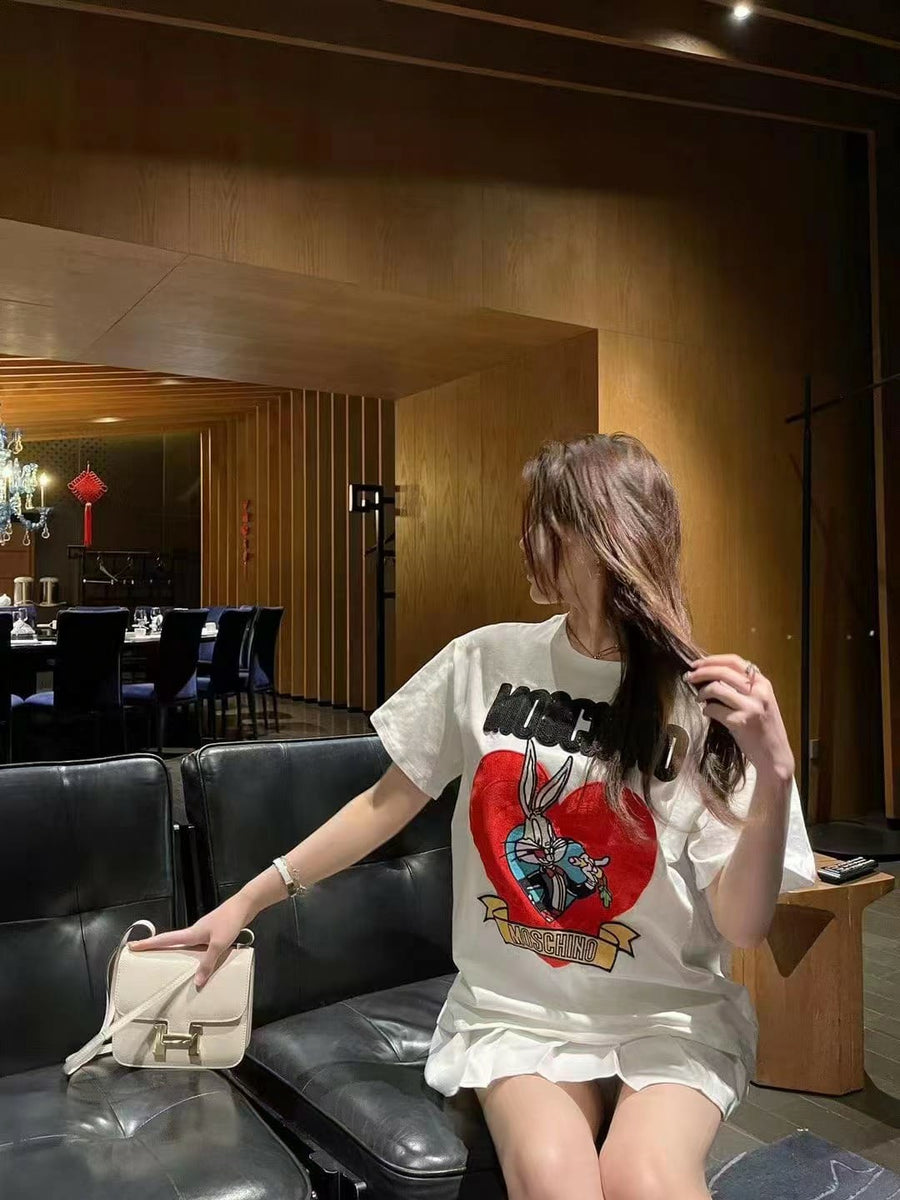 Woman in oversized white graphic T-shirt featuring Bugs Bunny in a red heart with Moschino banner, on a leather sofa.
