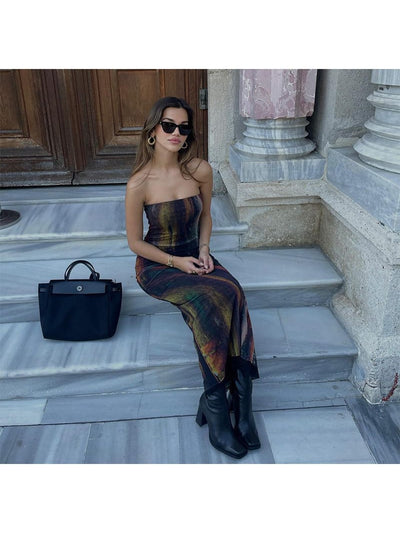 Woman in tie-dye mesh tube dress sits on marble steps, black boots, oversized sunglasses; black handbag beside.