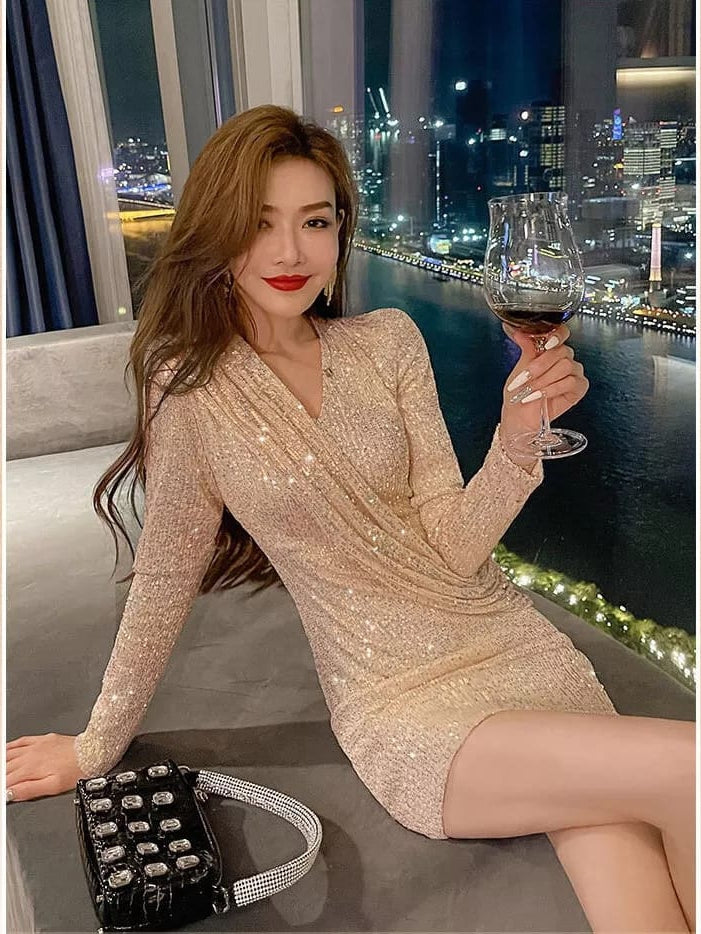 Woman in shimmering gold sequin wrap dress with long sleeves, red lipstick, holding wine glass by window at night.
